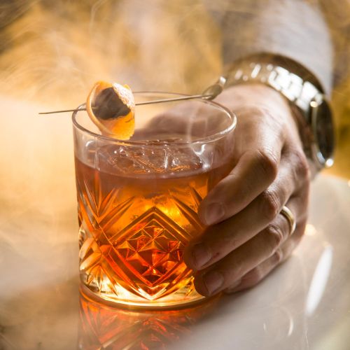 A hand holding a glass of amber whiskey with smoke or steam around it, warm lighting, close-up shot.