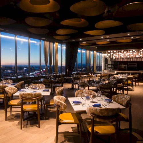 A stylish, warmly lit restaurant with wooden floors, round tables, yellow napkins, and a city view through large windows, cozy and upscale.