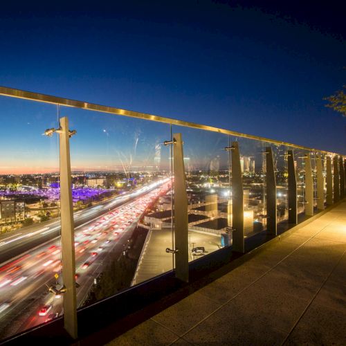 City lights along a waterfront boardwalk at night, with a railing and umbrella-lit tables under a clear evening sky.
