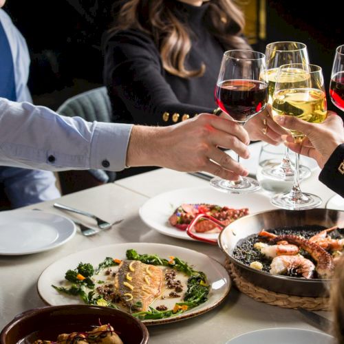 People eating a shared meal at a long table, plates of food in front, glasses, and people chatting in a cozy dining setting.